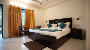 A bedroom with a large bed, a chair, a nightstand, and a view of the outdoors through curtains. - Wild Vista Corbett Resort (Dhikuli)