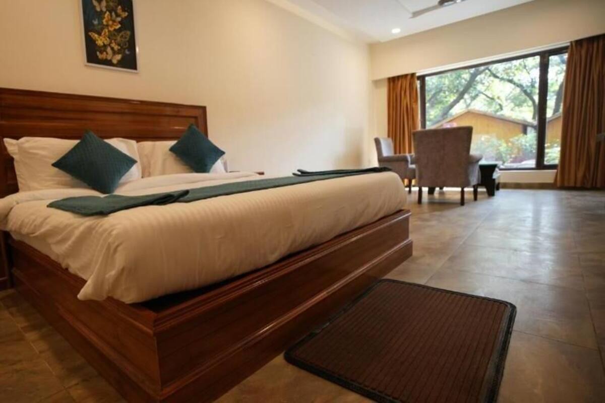 A hotel room with a large bed, a wooden headboard, a desk, and a chair. There is a window with curtains and a view of trees outside.