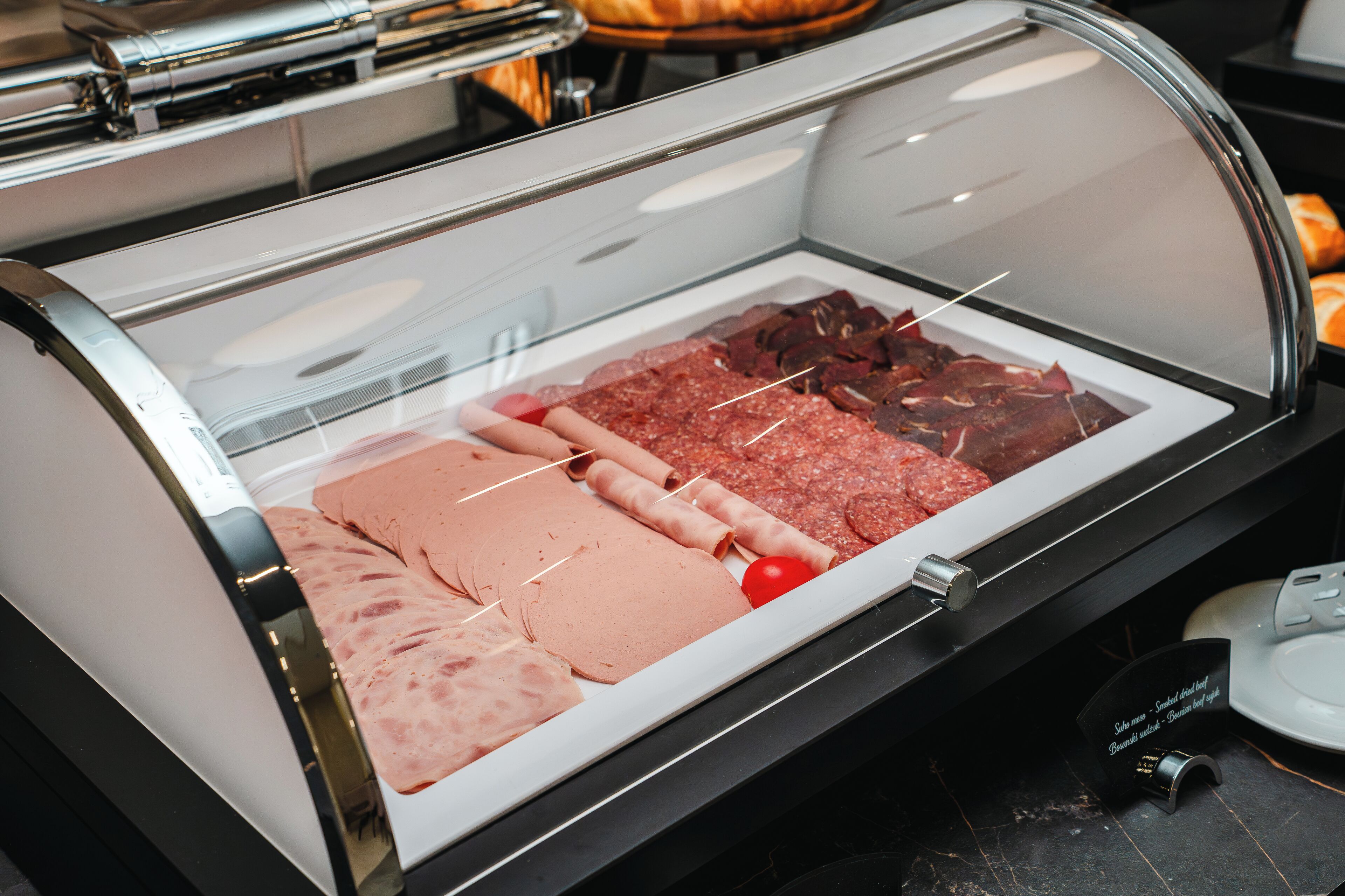 A meat display case with various types of sliced meats and a whole tomato.