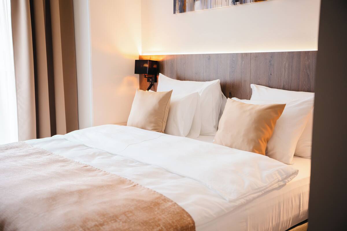 Una cama bien hecha con sábanas blancas y una manta beige, una lámpara de noche y un cabecero de madera.