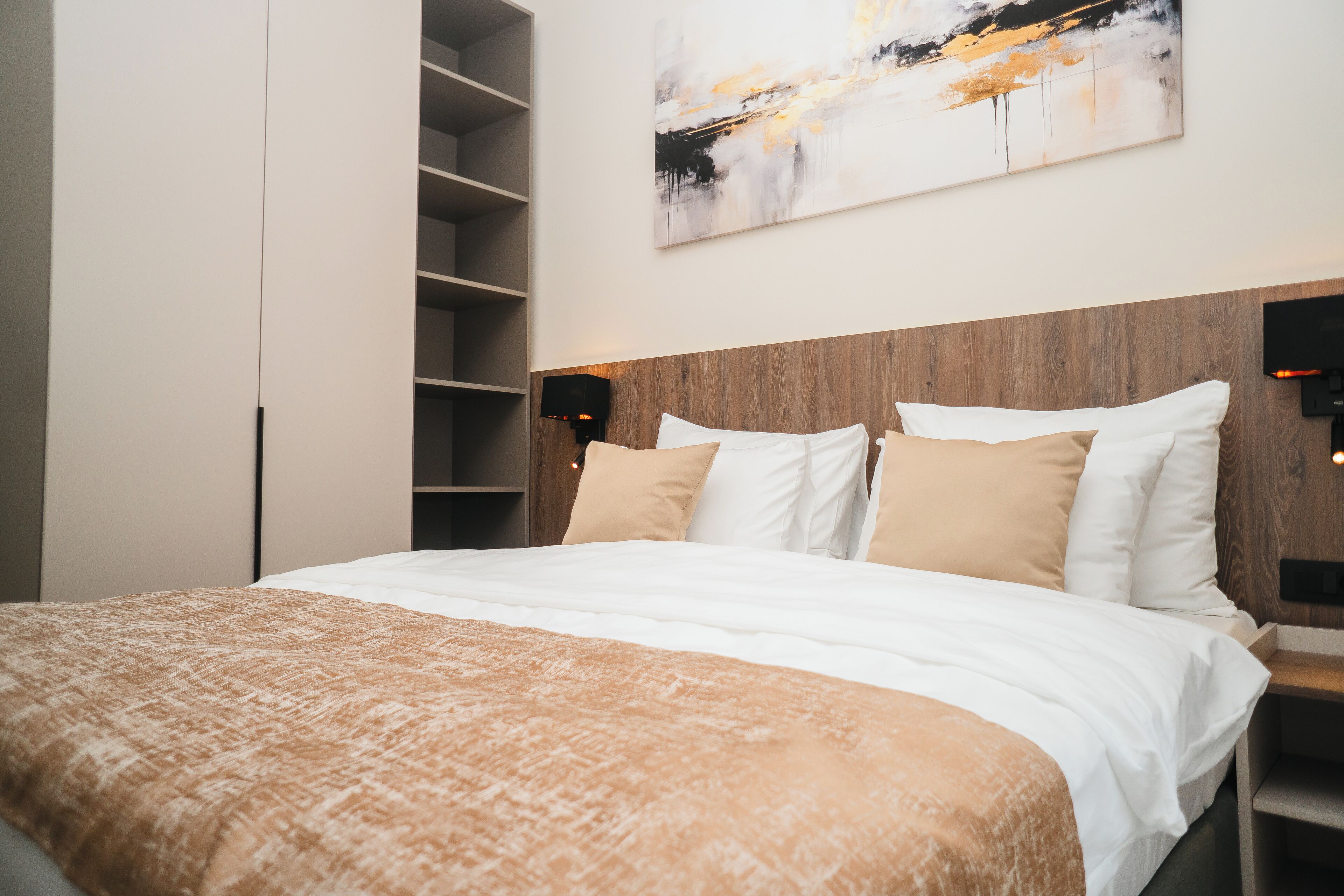 Ein modernes Schlafzimmer mit einem Holzkopfteil, einem großen Bett mit weißer und beiger Bettwäsche sowie Regalen an der linken Seite.