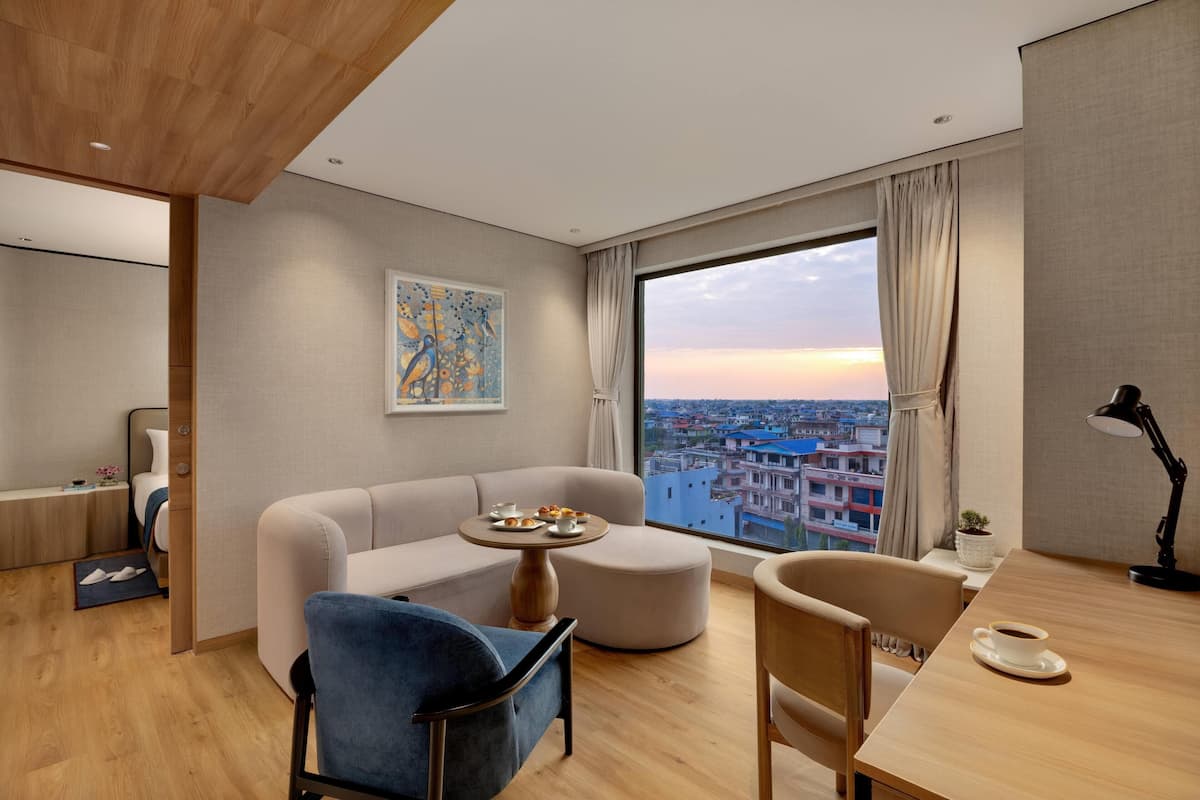 Une chambre d’hôtel moderne avec un canapé, une table à manger, une chaise, une petite table ronde sur laquelle se trouve une assiette de nourriture, une lampe et une grande fenêtre donnant sur la ville.