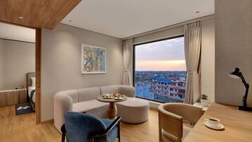 A modern hotel room with a sofa, a dining table, a chair, a small round table with a plate of food, a lamp, and a large window with a city view.