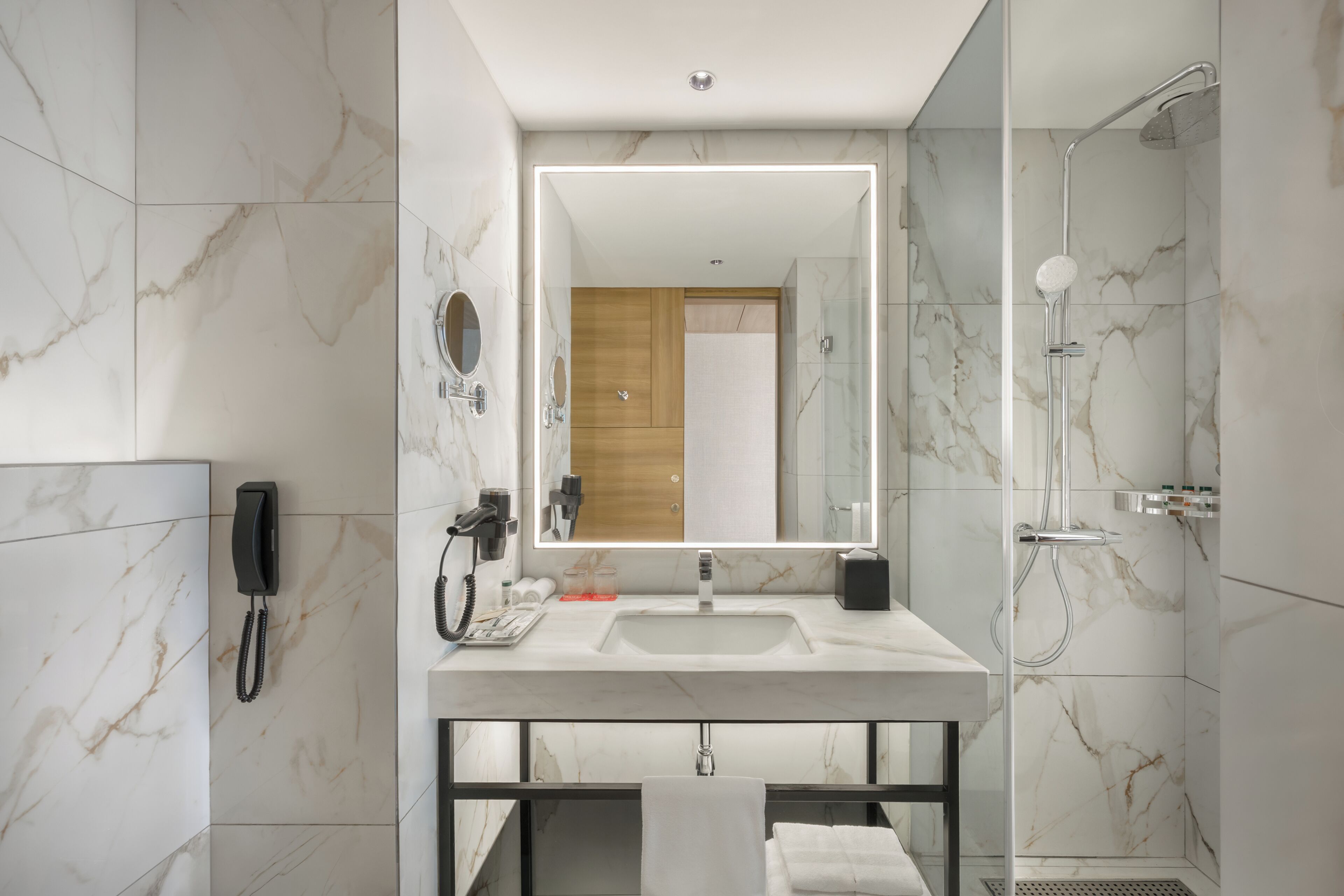 A modern bathroom with a marble-tiled wall, a large mirror, a sink, a handheld showerhead, and a telephone.