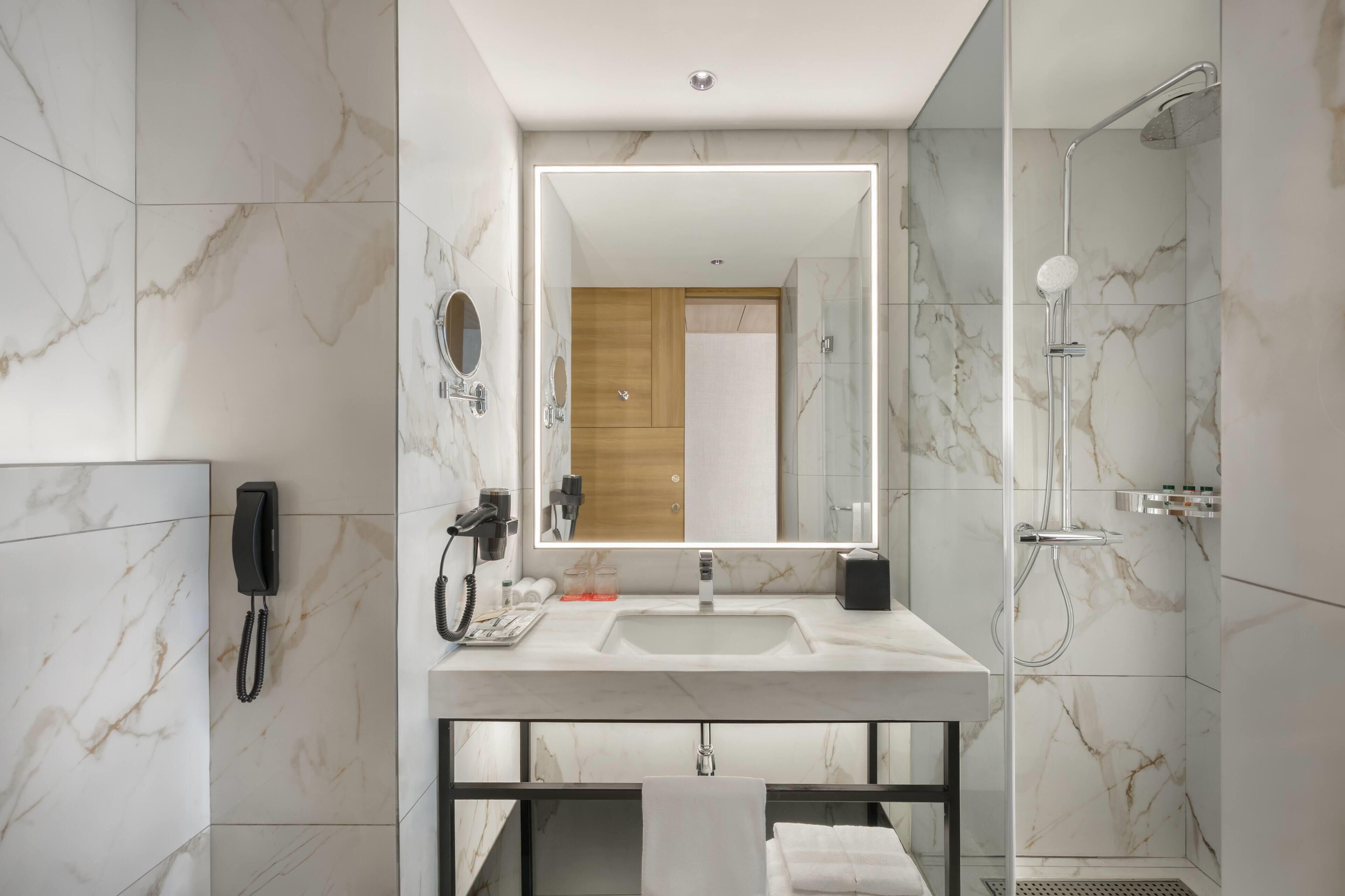 A modern bathroom with a marble-tiled wall, a large mirror, a built-in sink, a handheld showerhead, and a telephone.