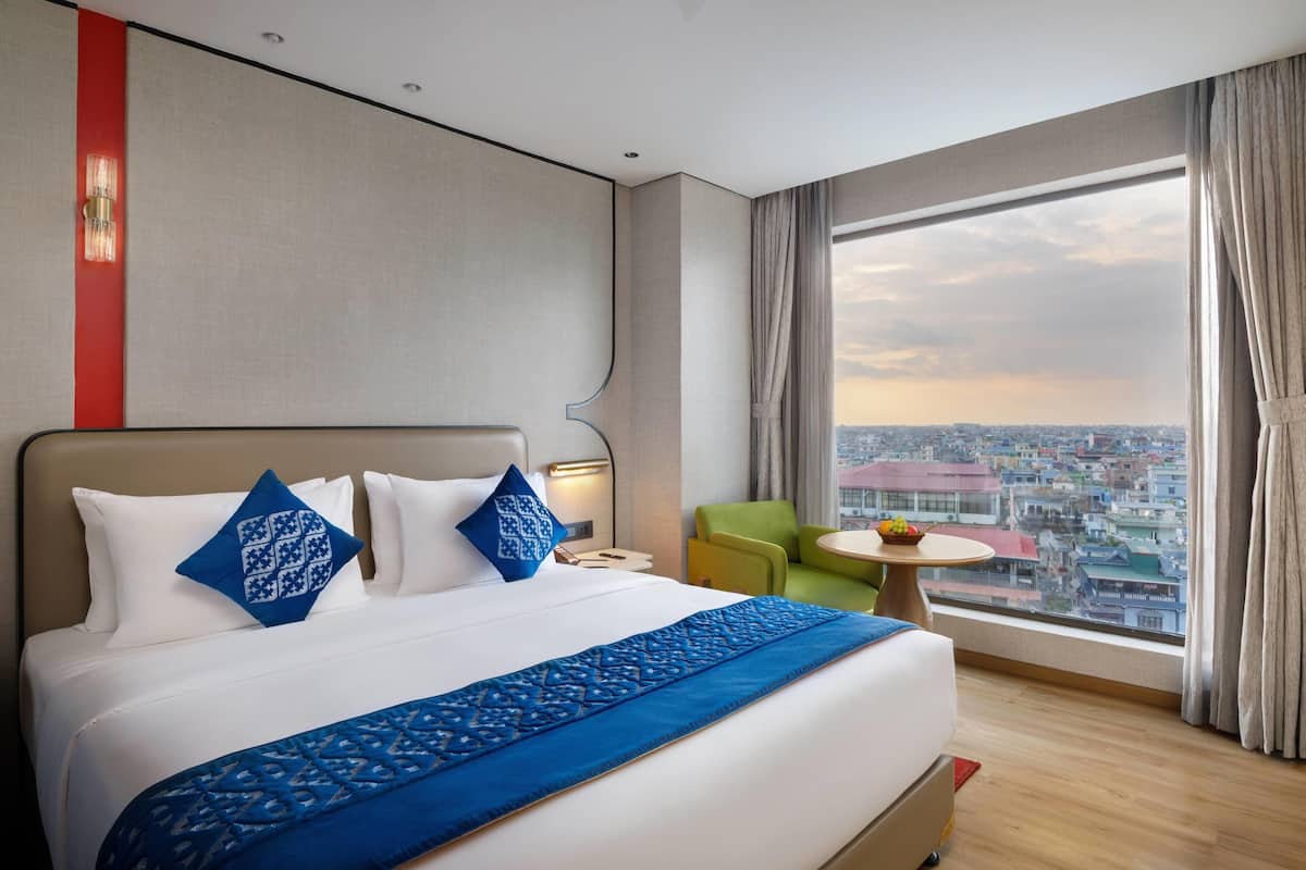 Une chambre d’hôtel avec un grand lit, une vue sur la ville, une petite table avec des fruits et un fauteuil vert.