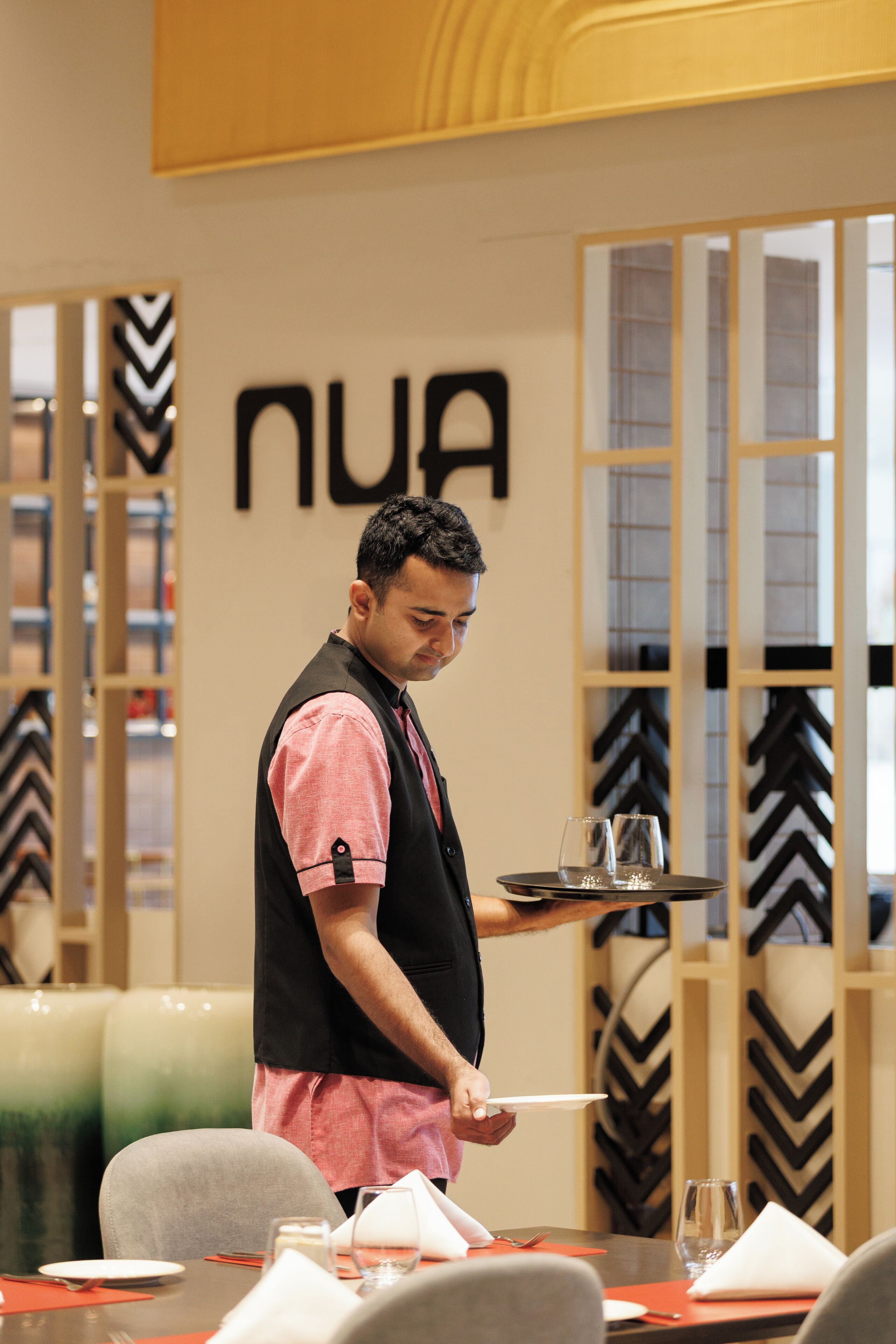 A person serving a tray with glasses and a plate in a modern dining area with a chevron-patterned wall and a visible 'NUA' sign.