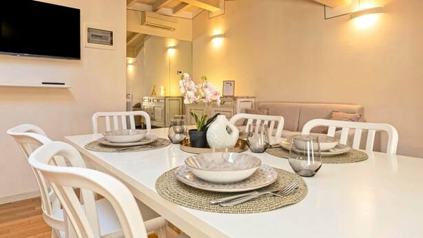 A dining area with a white table set for four, surrounded by white chairs. There is a television mounted on the wall above the table. - Family relaxation in downtown Bergamo (Bergamo)