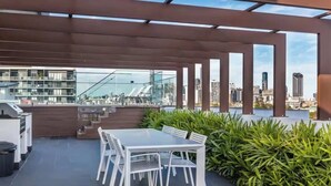 A rooftop terrace with a dining table and chairs, surrounded by greenery and overlooking a cityscape.