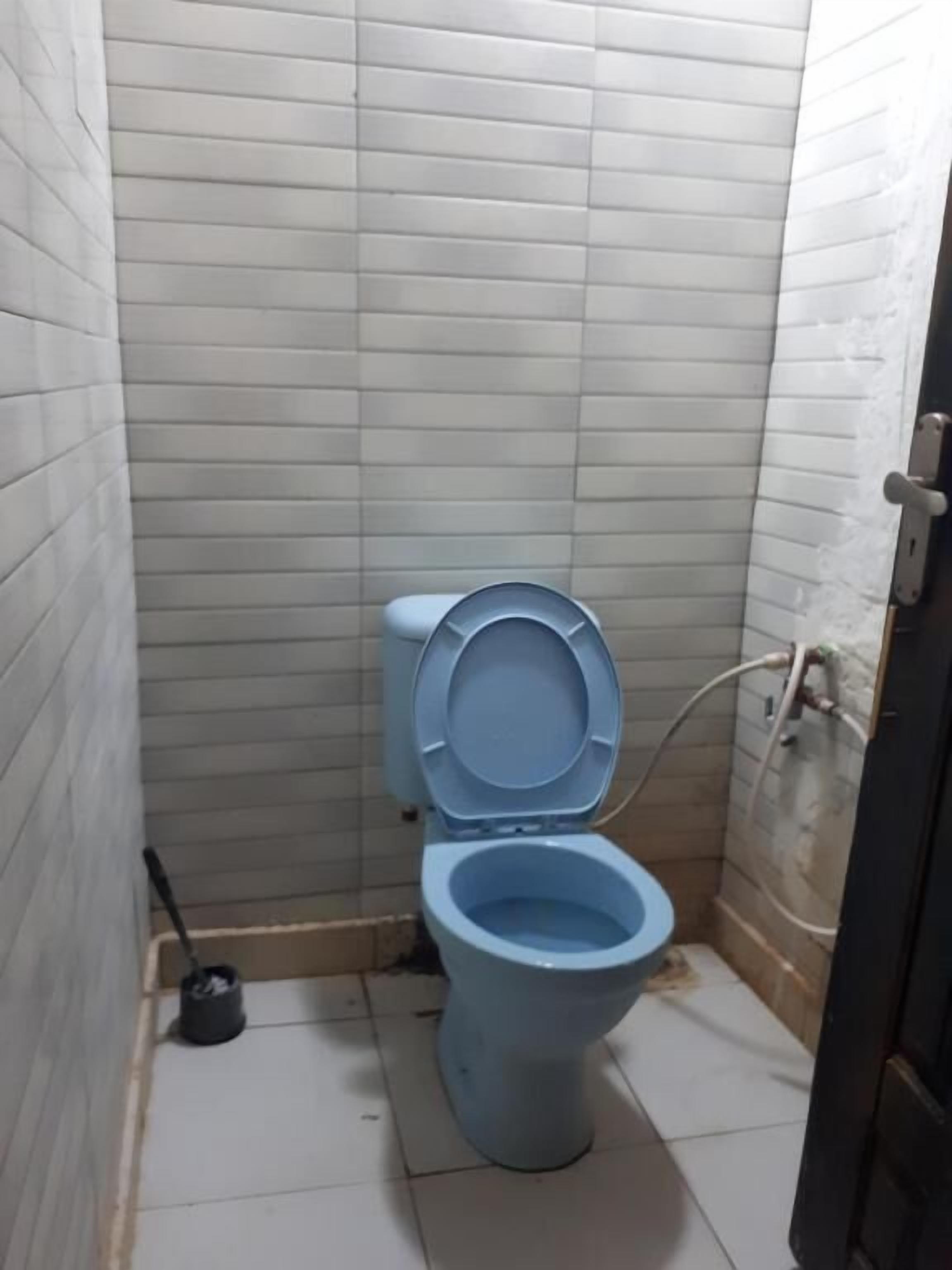 A small, narrow bathroom with a blue toilet, tiled walls, and a visible pipe on the right.