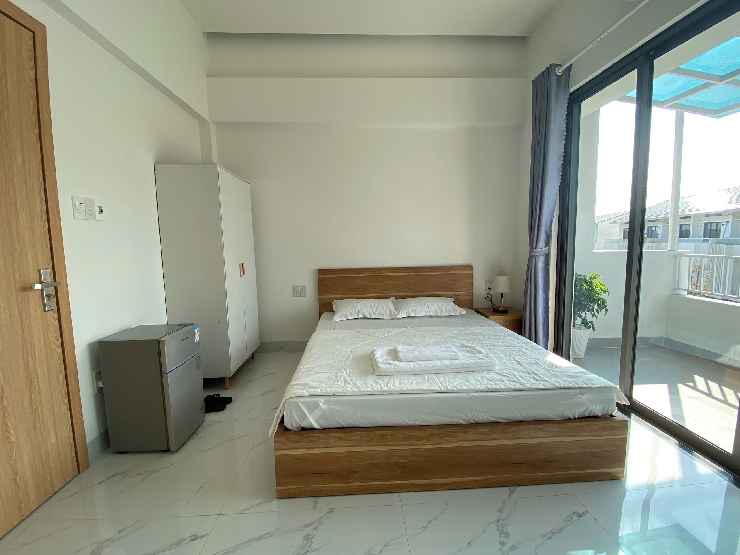 Een slaapkamer met een bed, een kledingkast, een kleine koelkast en een balkon met een plant.