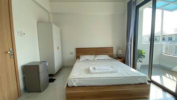 Een slaapkamer met een bed, een kledingkast, een kleine koelkast en een balkon met een plant.
