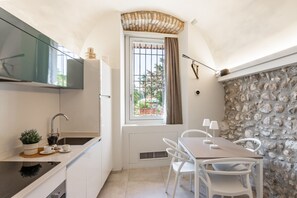A compact kitchen with a window, a stone wall, and a dining area with white chairs and a table. - Palazzo Trevignane - La Saletta Verde (San Felice del Benaco)