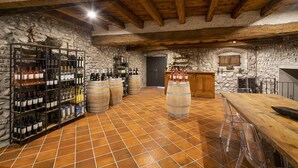 Una cantina rustica con pareti in pietra, travi in legno e pavimento piastrellato. Si notano scaffali per vini, botti e un'area bar con sgabelli.
