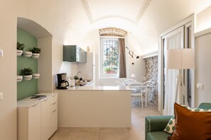 A compact kitchen with a central island, a dining area with chairs, a sofa, and a view through a window with a curtain. - Palazzo Trevignane - La Saletta Verde (San Felice del Benaco)