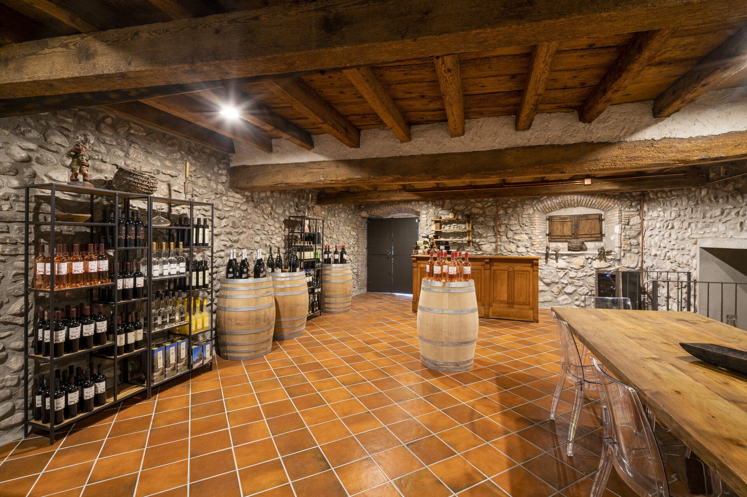 Ein rustikaler Keller mit Steinwänden, Holzbalken und Fliesenboden. Weinkisten, Fässern und eine Bar mit Hockern sind vorhanden.