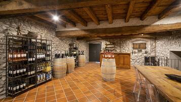 Ein rustikaler Keller mit Steinwänden, Holzbalken und Fliesenboden. Weinkisten, Fässern und eine Bar mit Hockern sind vorhanden.