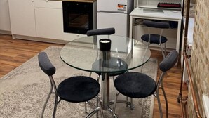 Kompakti keittiö, jossa on valkoiset kaapit, mikroaaltouuni, jääkaappi sekä lasinen ruokapöytä ja kaksi tuolia.