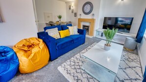 A living room with a blue sofa, a yellow bean bag, a glass coffee table, a TV, and a fireplace. - Modern 3-Bed House | Wednesbury | Garage + Driveway (Wednesbury)