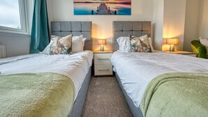 A hotel room with two beds, a nightstand with lamps, a painting of a sunset over water, and a view of the outdoors. - Modern 3-Bed House | Wednesbury | Garage + Driveway (Wednesbury)