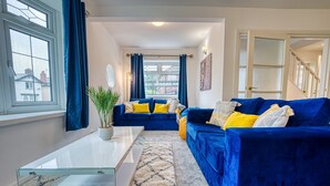 A living room with a blue sofa, a glass coffee table, and a view through a window with curtains. - Modern 3-Bed House | Wednesbury | Garage + Driveway (Wednesbury)