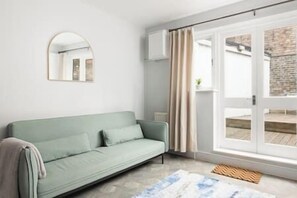 A room with a light green sofa, a mirror on the wall, a large window with curtains, and a view of an outdoor area with a brick wall. - New! Lovely 1BR Flat w/ Patio in Herne Hill (Greater London)
