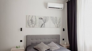 Et soveværelse med en grå polstret sengegavl, et gråt dynebetræk, to puder, en vægmonteret aircondition og et indrammet botanisk tryk.