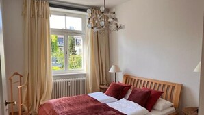 A bedroom with a wooden bed, beige curtains, a window with a view, and a hanging light fixture. - Seeliebe | city location | seeglueck (Konstanz)