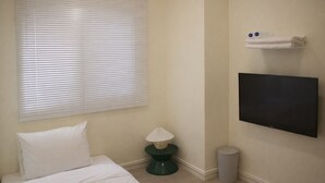 Quarto de hotel com cama, televisão fixada na parede, mesinha com abajur e lixeira.