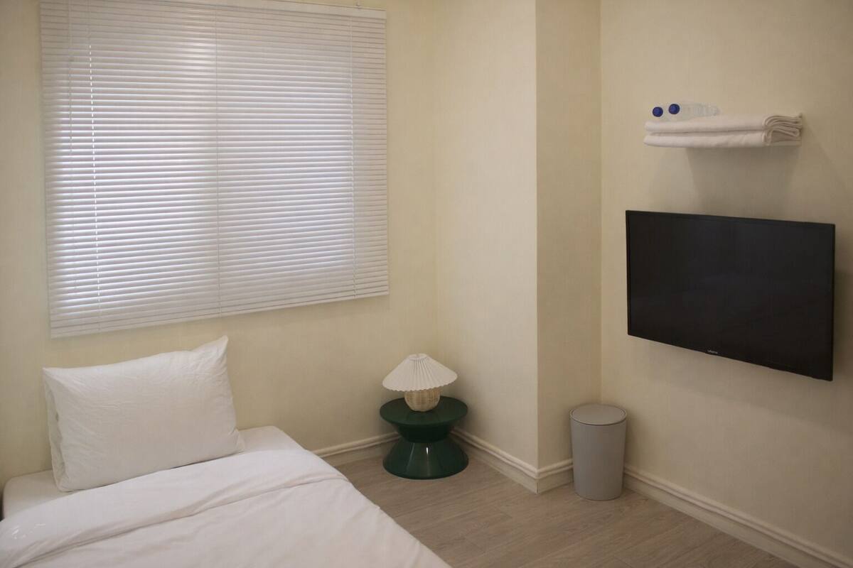 A hotel room with a bed, a TV mounted on the wall, a small table with a lamp, and a trash can.