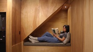 Uma pessoa sentada num banco, lendo um livro, em um ambiente com paredes de madeira e um banco embutido.