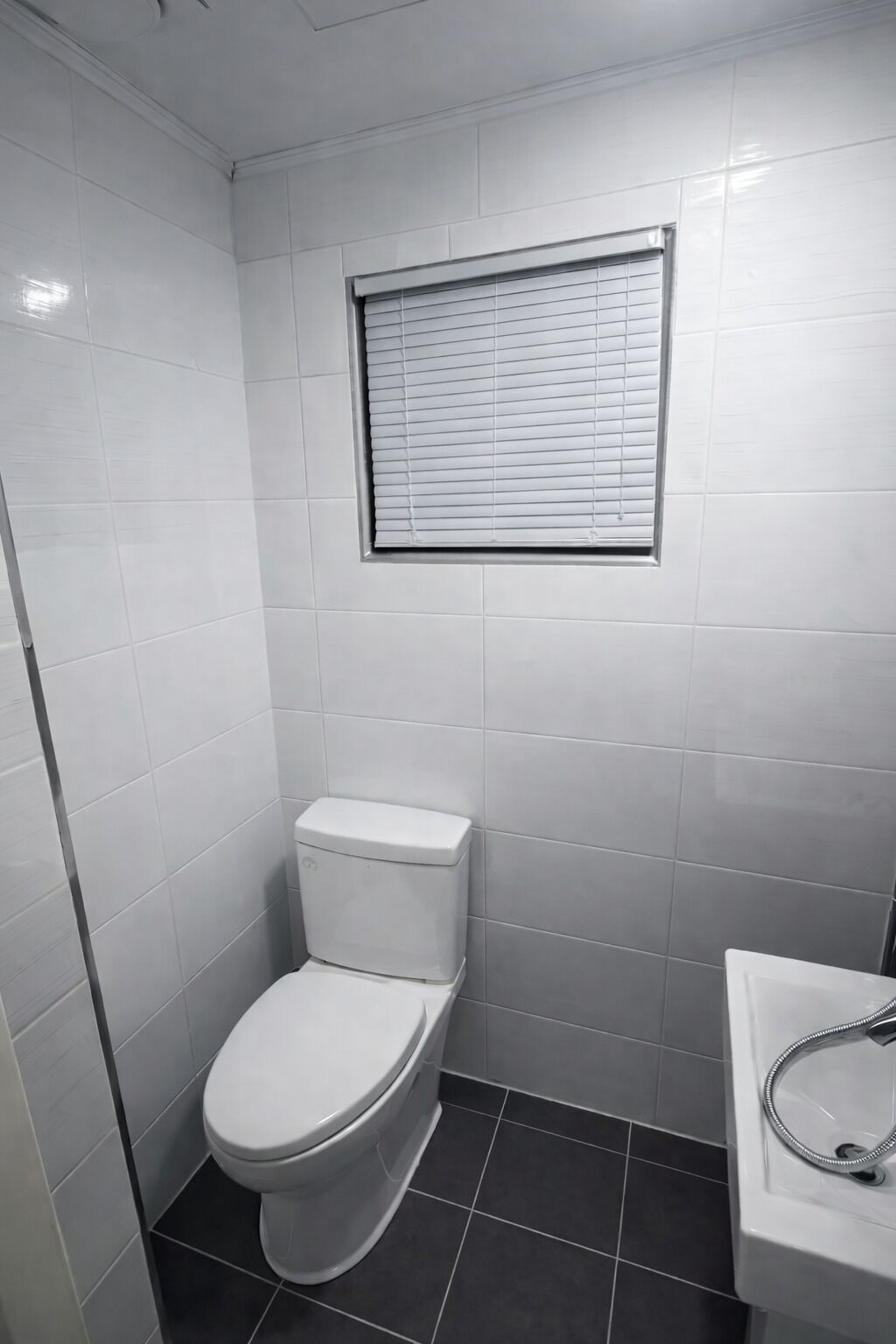 A small, modern bathroom with white tiles, a toilet, a window with blinds, and a sink with a chrome faucet.