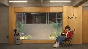 Um quarto com uma janela grande, uma divisória de madeira com a inscrição 'THE LOFT 6' e uma pessoa sentada em uma cadeira lendo um livro.