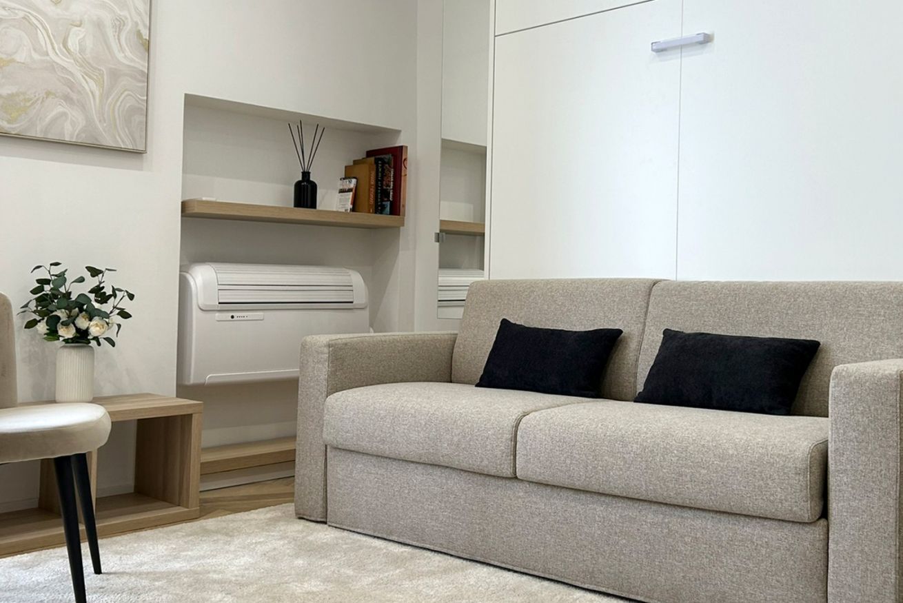 Un salon aux couleurs neutres, avec un canapé beige, une petite table d’appoint en bois et un radiateur blanc. Des étagères contenant des livres et des objets décoratifs sont fixées au mur.