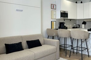 A modern living room with a beige sofa, two black cushions, and a kitchen area with white cabinets, a microwave, and a coffee machine. - Vatican Superior Suite (Roma)