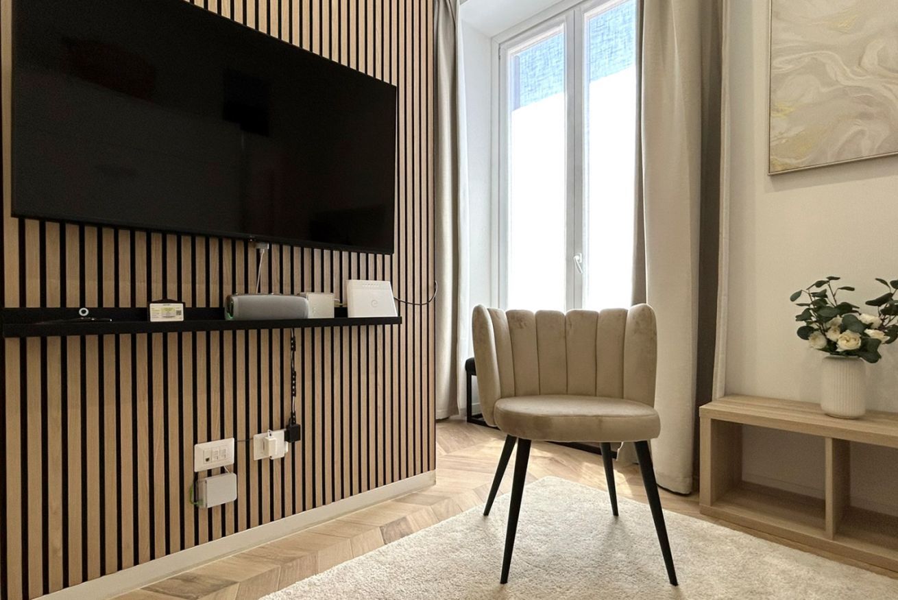 Uma sala de estar moderna com uma televisão de tela plana presa a uma parede de painéis de madeira, um sofá bege e uma mesa pequena com um vaso de flores.