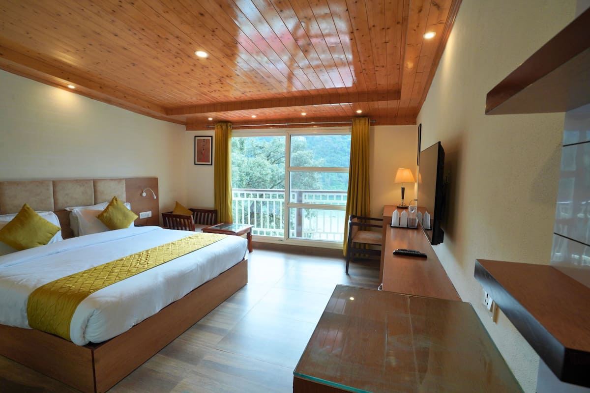 A hotel room with a wooden ceiling, a large bed with a yellow bedspread, a glass coffee table, a TV, and a balcony with a railing.