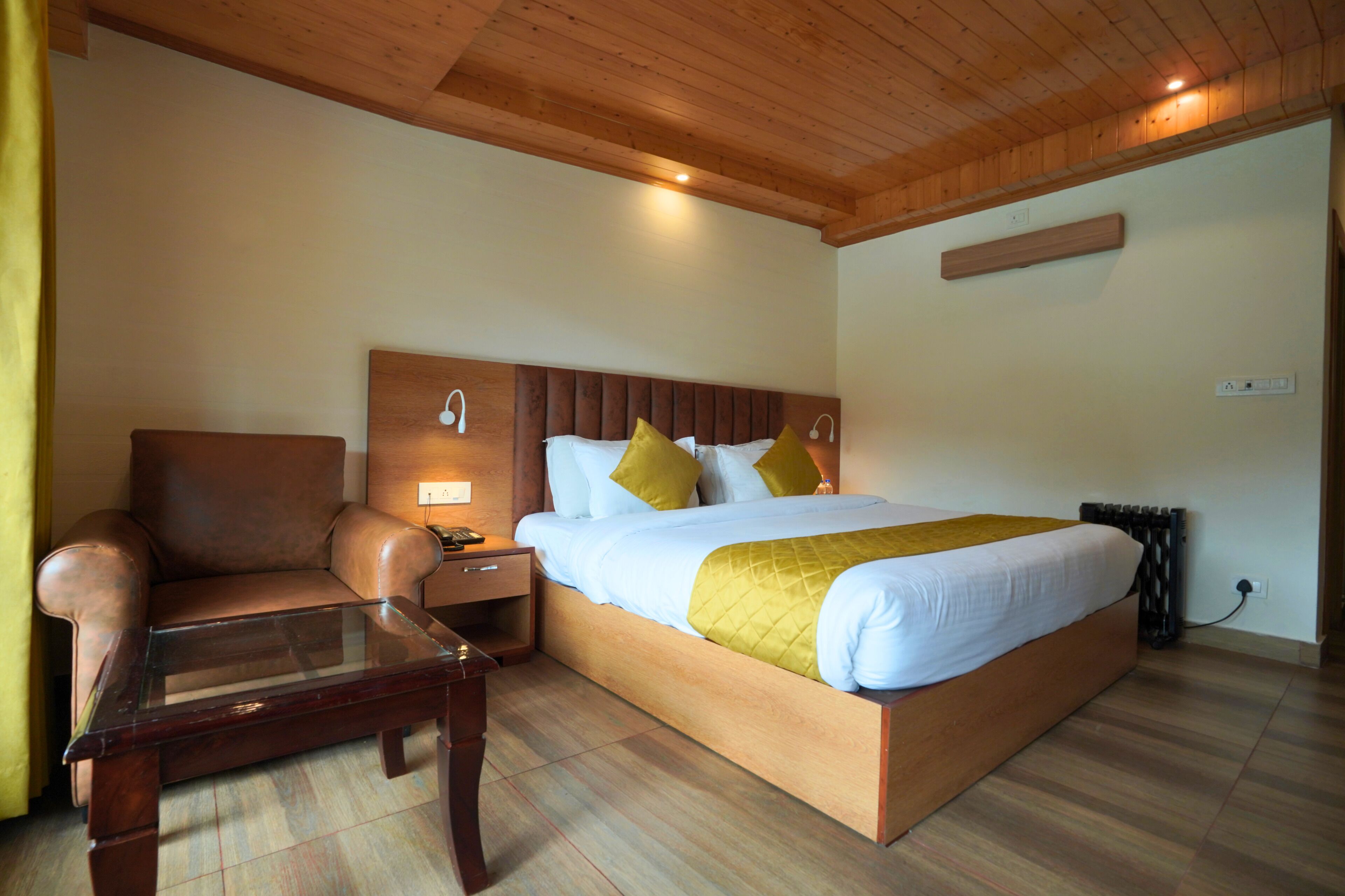 Un dormitorio con cabecera de madera, cama con ropa de cama blanca y amarilla, un sillón de cuero y una mesa con superficie de vidrio.