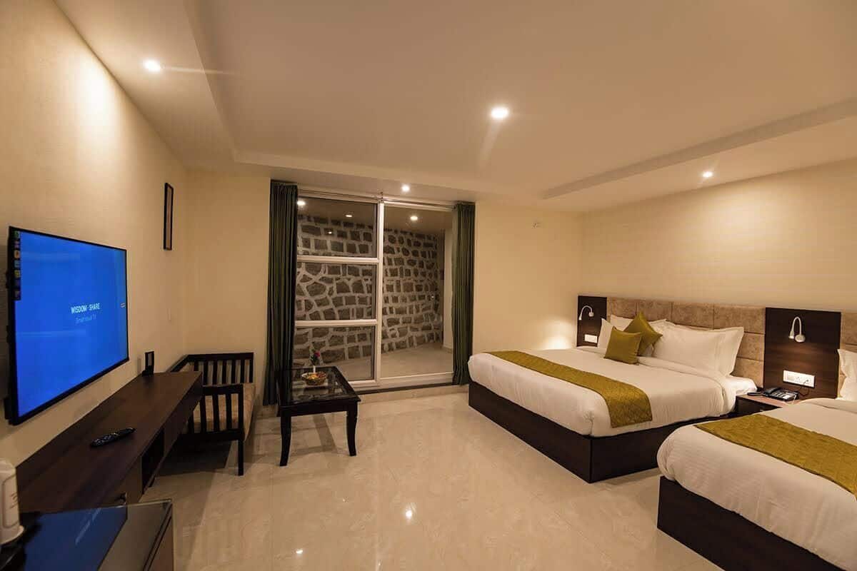 A hotel room with a TV, two beds, a desk, and a sliding glass door to a stone wall.