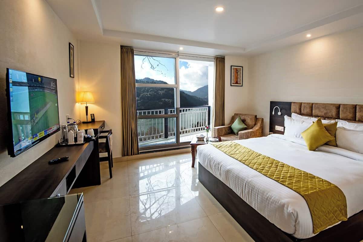 A hotel room with a bed, a TV, a desk, a chair, and a balcony with a mountain view.