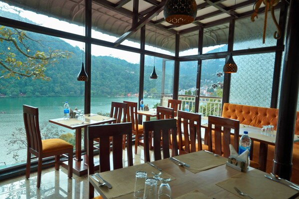 A restaurant with tables and chairs, a view of a lake, and hanging light fixtures. - The Imperial Green (180 lake view) (Nainital)