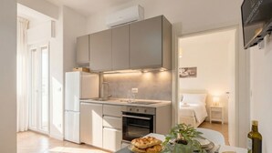 Eine moderne Küche mit Einbauofen, eine Essecke mit Tisch für zwei Personen und ein Schlafzimmer mit Bett und Nachttisch.