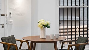 A room with a round wooden table, two chairs, a vase with flowers, and a view through a barred window. - Laneway Living | 1BR Heritage Stay in CBD (Melbourne)