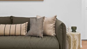 A living room with a sofa, pillows, a coffee table with books and cups, a painting on the wall, and a small side table with a candle. - Laneway Living | 1BR Heritage Stay in CBD (Melbourne)