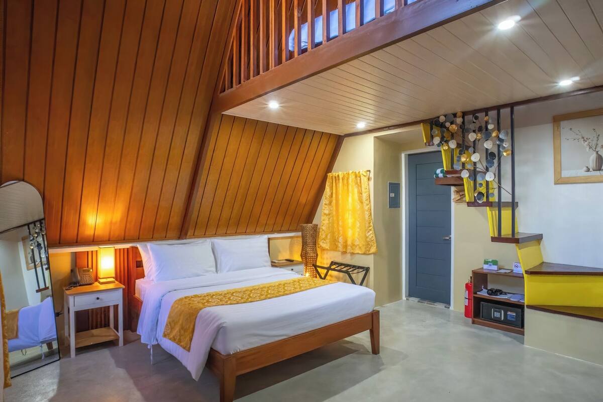 Une chambre avec un plafond en bois, un lit, une table de chevet, une lampe, une porte et une étagère fixée au mur, ornée d’objets décoratifs.