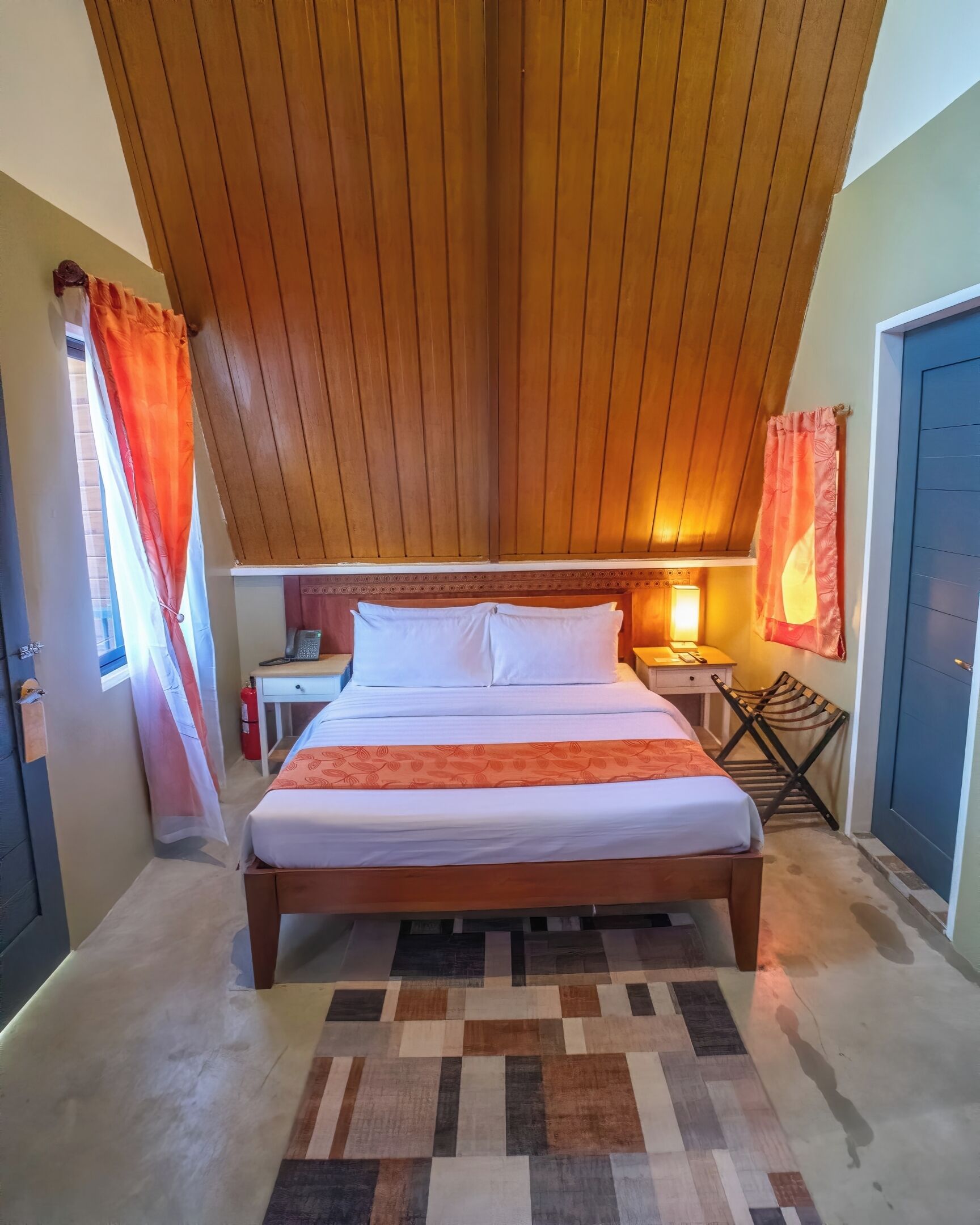 Ein Schlafzimmer mit Holzbalkendecke, einem Bett mit weißer Bettwäsche, einem Nachttisch, einer Lampe und einem Fenster mit orangefarbenen Vorhängen.