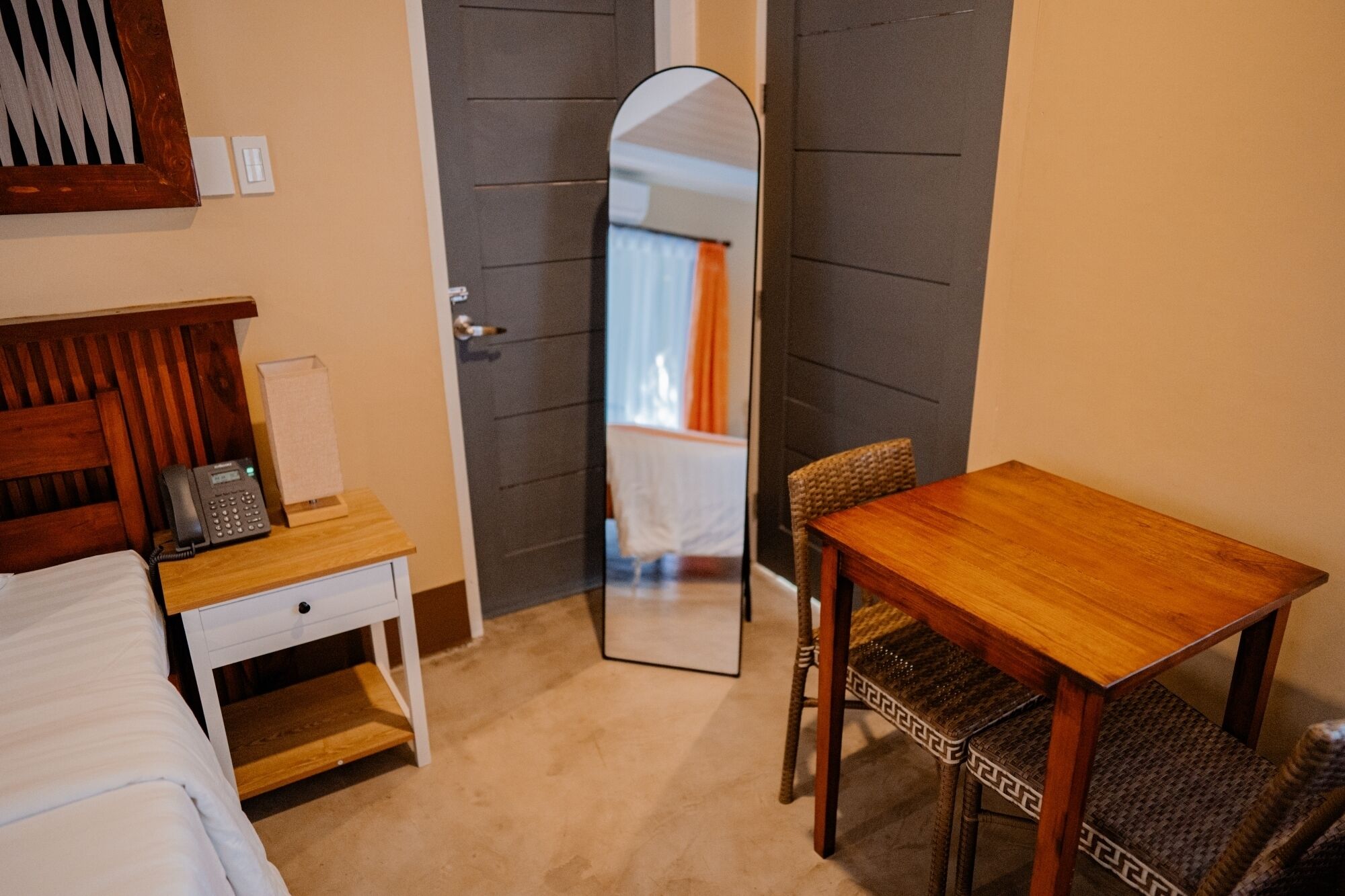 A room with a bed, bedside table with a phone, a wooden chair, a small table, and a mirror.