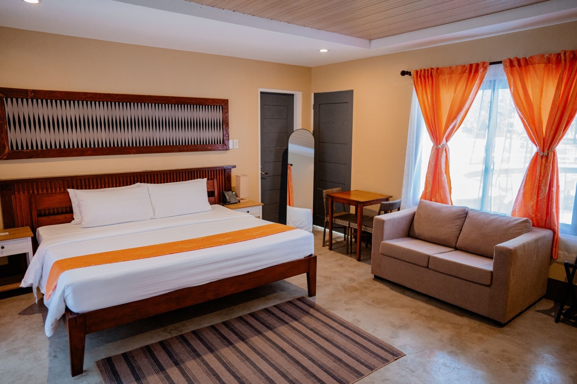 Habitación de hotel con cama, sofá, una mesita, espejo y ventana con cortinas.