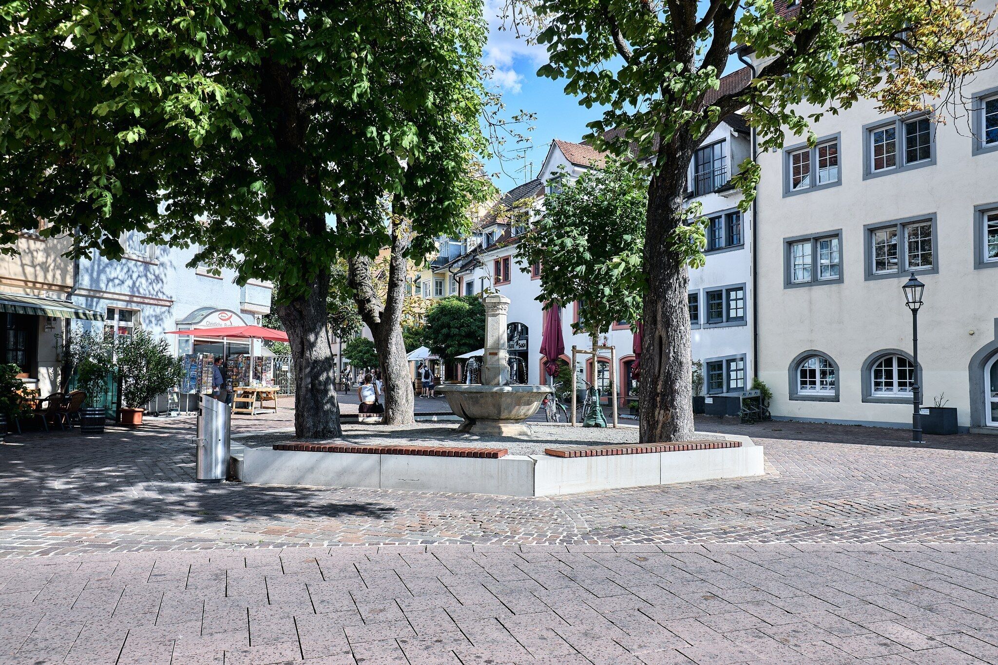 A cobblestone square with a central fountain, surrounded by trees and buildings with traditional architecture.