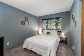 A bedroom with a large bed, two bedside tables with lamps, a window with blinds, and three framed pictures on the wall.